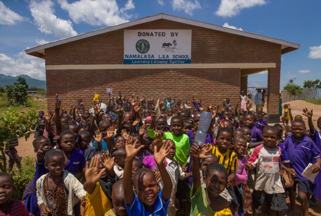 Young students gathered outside the Namalasa LEA School brick building with a sign which designates the school was donated by African Parks and Liwonde National Park with their corresponding logos. The school's motto "Learning & Growing Together" appears below its name.